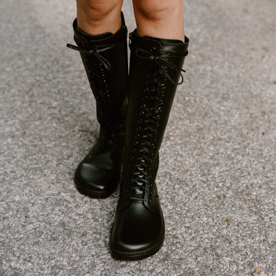 #A photo of Peerko Empire boots made from leather and rubber soles. The boots are black in color, they are a tall riding boot style with laces all the way up and a side zipper. Both boots are shown from the front and worn on a woman's feet with a view of her knees down. Her knees are bare, and she is standing on a paved road with her left foot slightly in front of her right. #color_black