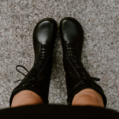 #A photo of Peerko Empire boots made from leather and rubber soles. The boots are black in color, they are a tall riding boot style with laces all the way up and a side zipper. Both boots are shown from above on a woman's feet, with view of the the bottom of a woman's black dress and bare knees down.  She is standing on a paved road. #color_black