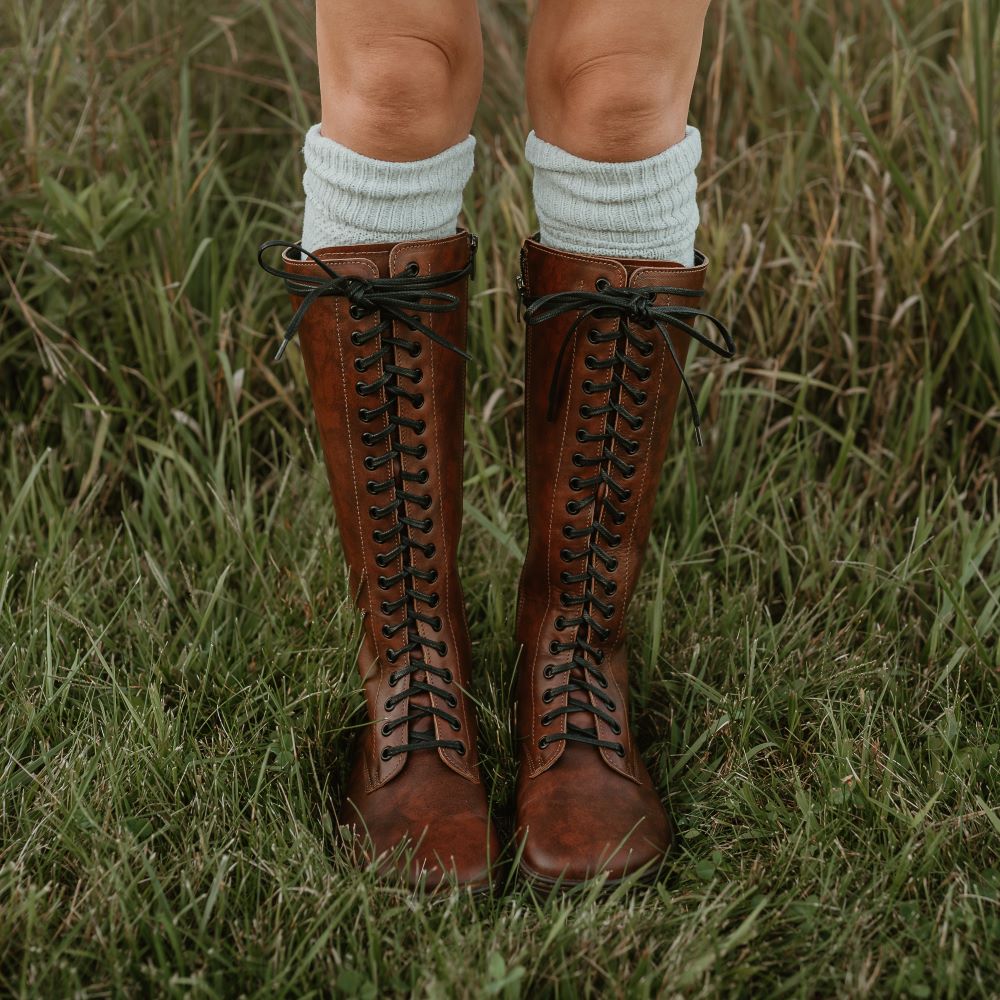 A photo of Brandy Peerko Empire tall leather riding boots with black laces all the way up, a side zipper, and black rubber soles. Both shoe are shown from the front with on a woman wearing cream socks peeking out above the boots standing in grass. #color_brandy