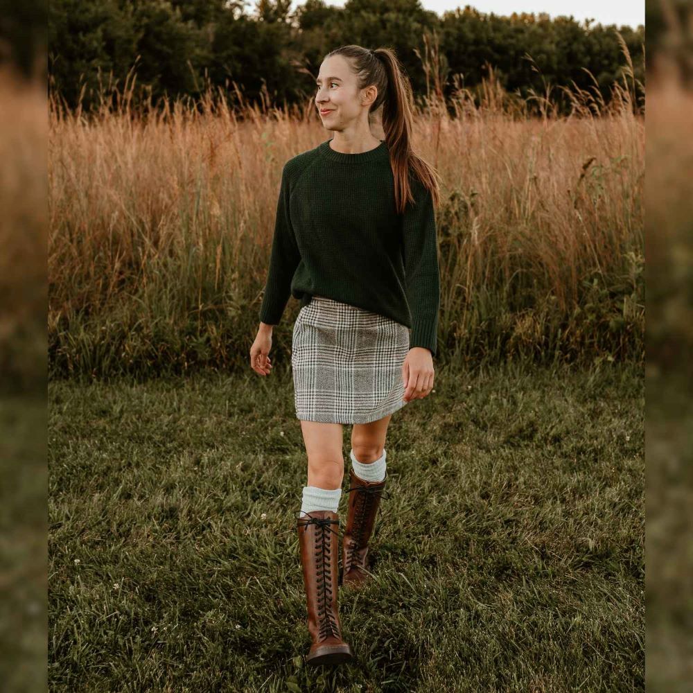 A photo of Brandy Peerko Empire tall leather riding boots with black laces all the way up, a side zipper, and black rubber soles. Both shoe are shown from the front on a woman wearing a cream plaid skirt and dark green sweater with cream socks peeking out above the boots. She is walking towards the camera standing in grass with tall grass and trees behind. #color_brandy