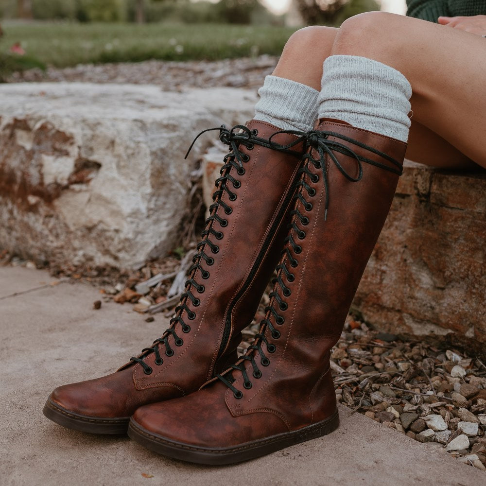 A photo of Brandy Peerko Empire tall leather riding boots with black laces all the way up, a side zipper, and black rubber soles. Both shoe are shown facing left on a woman wearing a cream plaid skirt and dark green sweater with cream socks peeking out above the boots. She is sitting on a rock retaining wall next to a sidewalk with grass and trees behind. #color_brandy