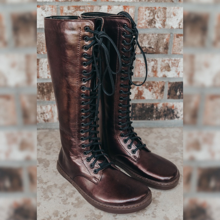 A photo of Peerko Empire boots made from leather and rubber soles. The boots are chestnut metallic in color, they are a tall riding boot style with laces all the way up and a side zipper. Both boots are shown diagonally from the front right against a brick background. #color_chestnut