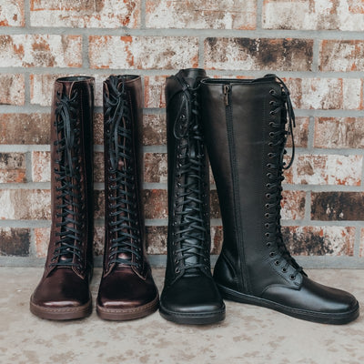 A photo of Peerko Empire boots made from leather and rubber soles. Two pairs of tall riding boot style with laces all the way up and a side zipper are shown here. Chestnut colored boots are shown from the front on the left. Black boots are shown with thr right boot facing front and the left boot pointing to the right.  Shoes are shown against a brick wall background. #color_chestnut