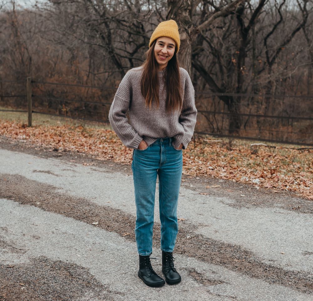 A photo of Peerko Frost combat boots made with smooth leather, wool, and rubber soles. The boots are black in color, wool lined, with silver speed hooks at the top. Both boots are shown from the front on a woman's feet. The woman is wearing blue straight leg jeans and a brown sweater with a yellow beanie hate and is standing with her hands in her pockets on a paved road #color_black