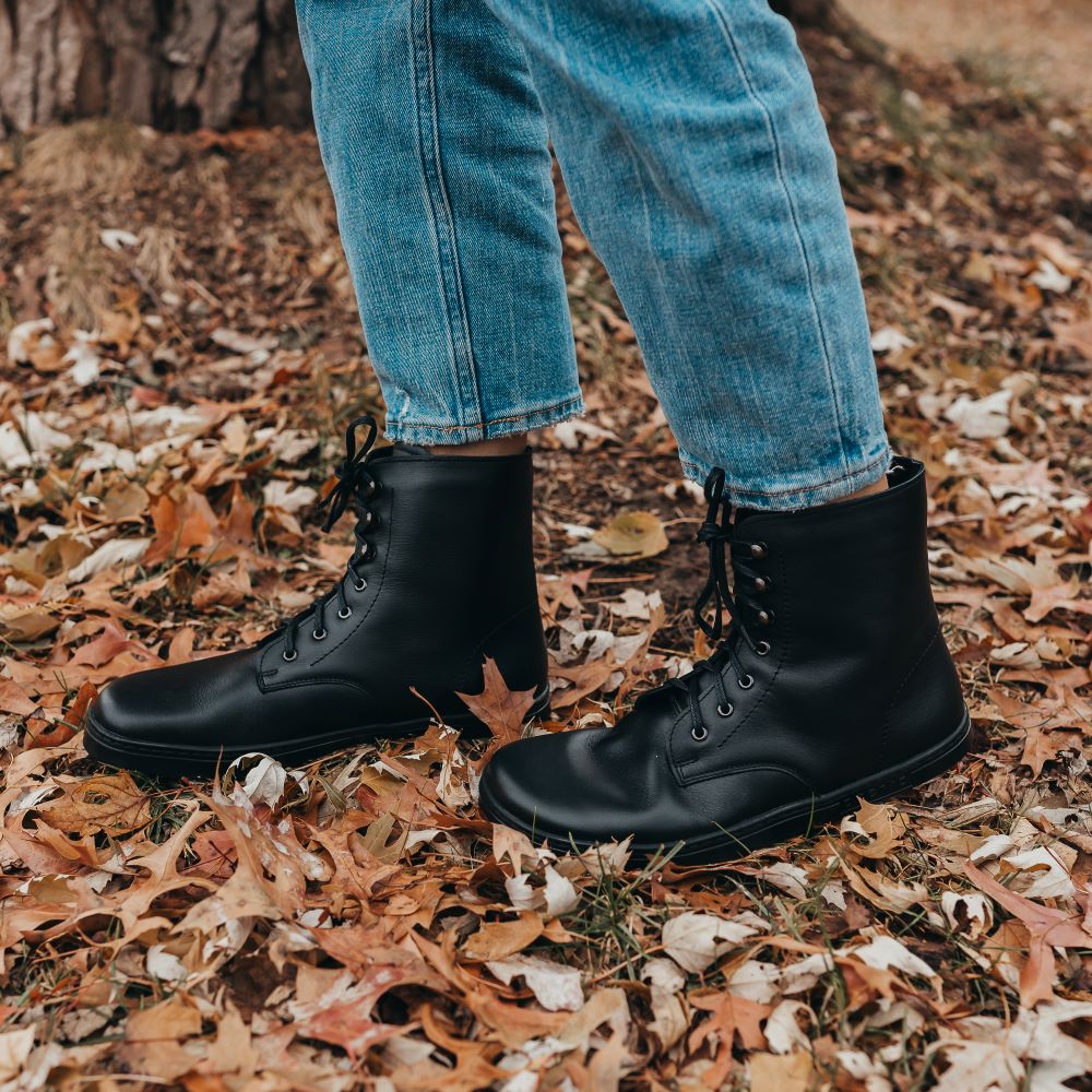 A photo of Peerko Frost combat boots made with smooth leather, wool, and rubber soles. The boots are black in color, wool lined, with silver speed hooks at the top. Both boots are shown from the left side on a woman's feet with a view of her knees down. The woman is wearing blue straight leg jeans and is standing on fallen leaves with her right foot in front of her left. #color_black