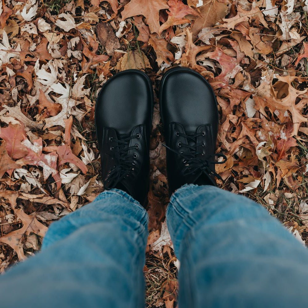 A photo of Peerko Frost combat boots made with smooth leather, wool, and rubber soles. The boots are black in color, wool lined, with silver speed hooks at the top. Both boots are shown from above on a woman's feet with a view of her knees down. The woman is wearing blue straight leg jeans and is standing on fallen leaves. #color_black