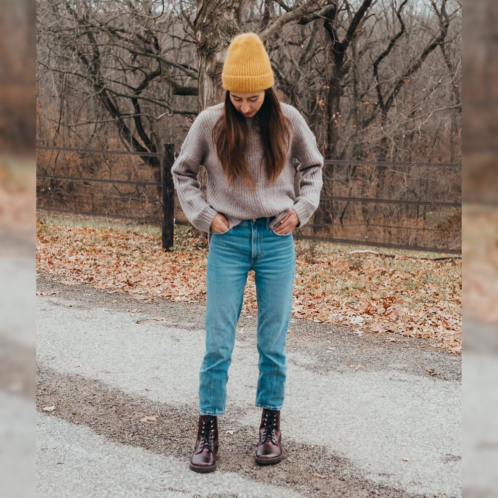 A photo of Peerko Frost combat boots made with smooth leather, wool, and rubber soles. The boots are chestnut metallic in color, wool lined, with silver speed hooks at the top. Both boots are shown from the front on a tan woman with long brown hair wearing light-wash cropped blue jeans and a loose grey knit sweater and a fizzy yellow knit beanie. Woman is standing on a grey paved road with a fall woods scene in the background. #color_chestnut