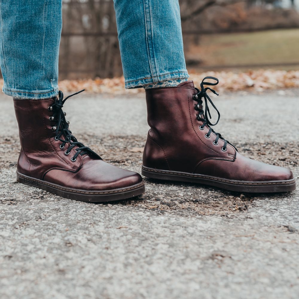 A photo of Peerko Frost combat boots made with smooth leather, wool, and rubber soles. The boots are chestnut metallic in color, wool lined, with silver speed hooks at the top. Both boots are shown facing right with the left foot in front of the right on a woman standing on a grey paved road wearing light-wash cropped blue jeans with trees and grass in the background. #color_chestnut