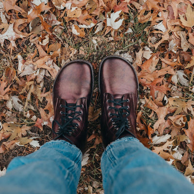 A photo of Peerko Frost combat boots made with smooth leather, wool, and rubber soles. The boots are chestnut metallic in color, wool lined, with silver speed hooks at the top. Both boots are shown ftom above on a woman wearing light-wash cropped blue jeans standing on leaf covered grass. #color_chestnut