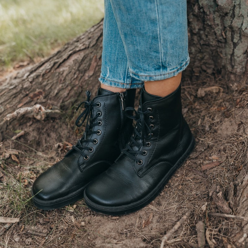 A photo of Peerko Go combat boots made with smooth leather and rubber soles. The boots are black in color, fleece lined, with a zipper at the side. Both boots are shown diagonally from the left on a womans feet standing next to a tree trunk and roots. #color_black