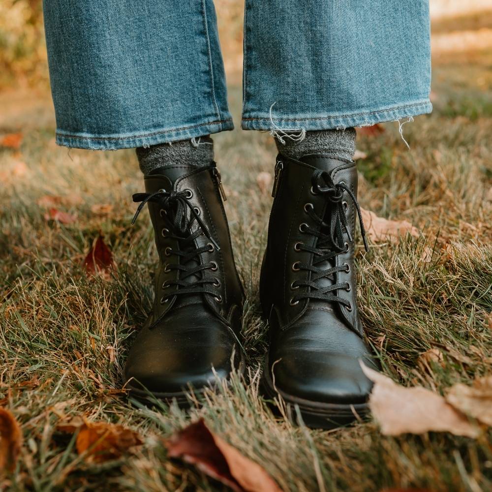 Black leather Peerko Go combat boots made with fleece lining, a black zipper, black laces, and black rubber soles. Both boots are shown from the front on a woman wearing cropped, loose, light-wash jeans and grey socks standing on leafy grass. #color_black