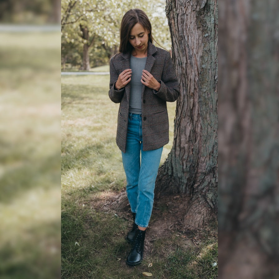 A photo of Peerko Go combat boots made with smooth leather and rubber soles. The boots are black in color, fleece lined, with a zipper at the side. Both boots are shown from the front on a womans feet standing in grass next to a tree trunk and roots. Woman is wearing loose blue jeans, a grey shirt, and a long, plaid blazer. #color_black