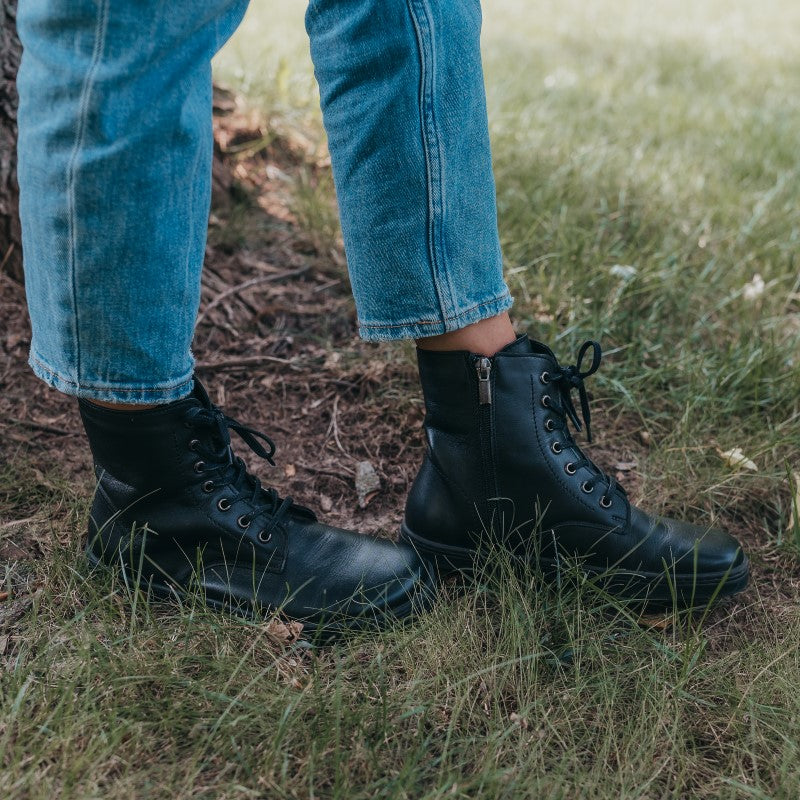 A photo of Peerko Go combat boots made with smooth leather and rubber soles. The boots are black in color, fleece lined, with a zipper at the side. Both boots are shown from the right on a womans feet standing in grass next to a tree trunk and roots. Woman is wearing loose blue jeans. #color_black