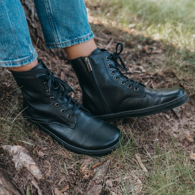 A photo of Peerko Go combat boots made with smooth leather and rubber soles. The boots are black in color, fleece lined, with a zipper at the side. Both boots are shown diagonally from the front right on a womans feet standing next to a tree trunk and roots. #color_black