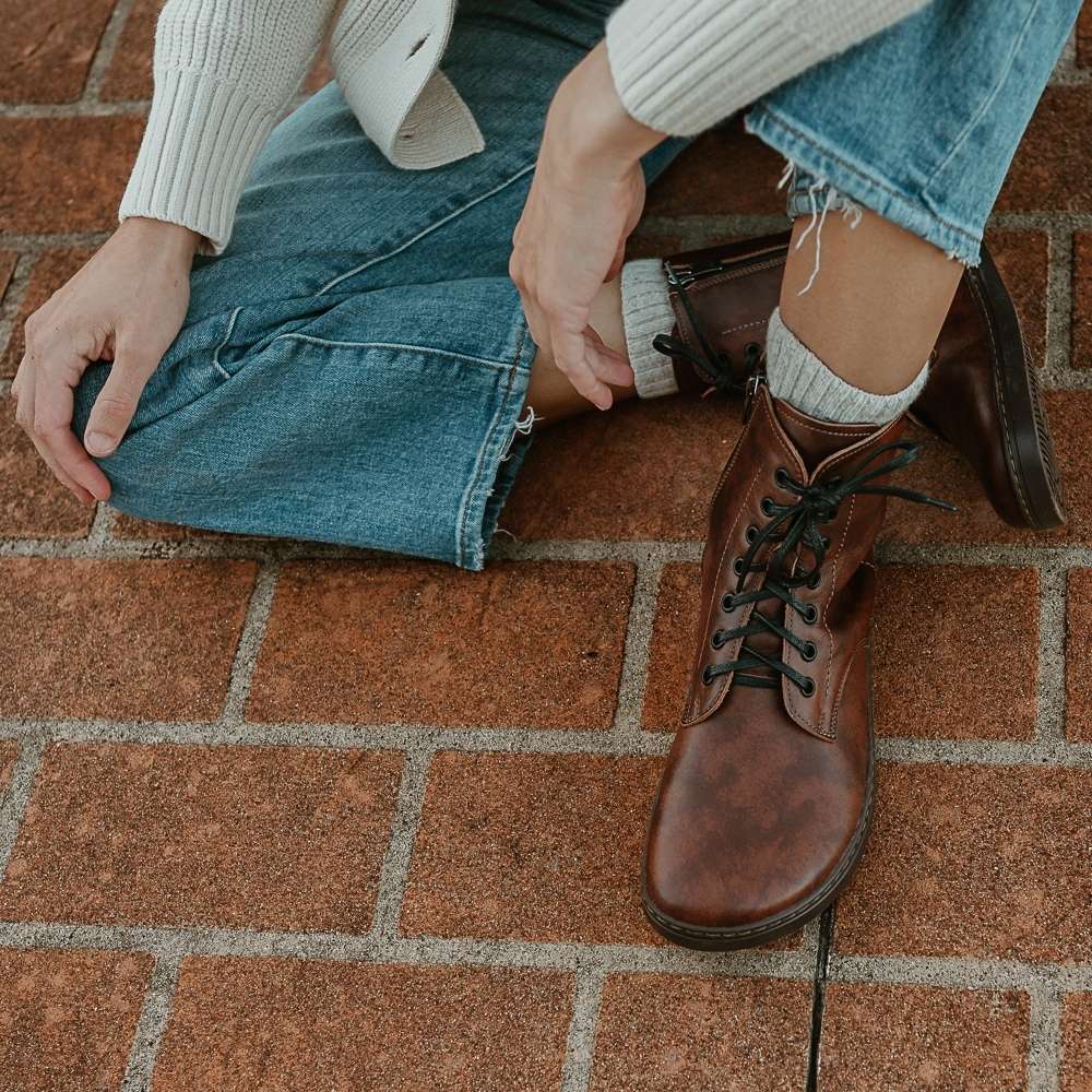 Brandy leather Peerko Go combat boots made with fleece lining, a black zipper, and black rubber soles. Both shoes are shown from above on a light-skinned woman sitting on a brick path with one leg crossed under the other wearing loose, distressed, light-wash jeans, a cream sweater, and a cream, knit cardigan. #color_brandy