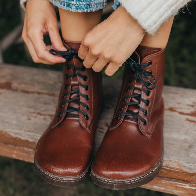 A photo of Peerko Go combat boots made with smooth leather, inside side zippers, and rubber soles. The boots are brown in color and fleece lined. Both boots are shown from the front on a womans feet wearing loose blue jeans and a fuzzy white sweater sitting on an old picnic table. #color_cognac
