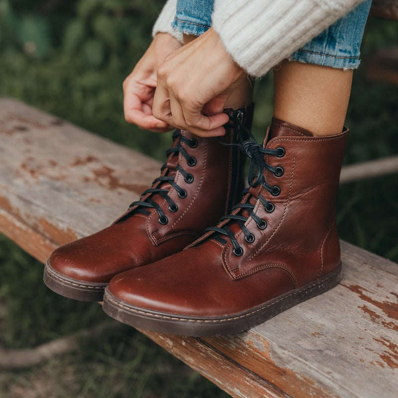A photo of Peerko Go combat boots made with smooth leather, inside side zippers, and rubber soles. The boots are brown in color and fleece lined. Both boots are shown diagonally from the front left on a womans feet wearing loose blue jeans and a fuzzy white sweater sitting on an old picnic table. #color_cognac