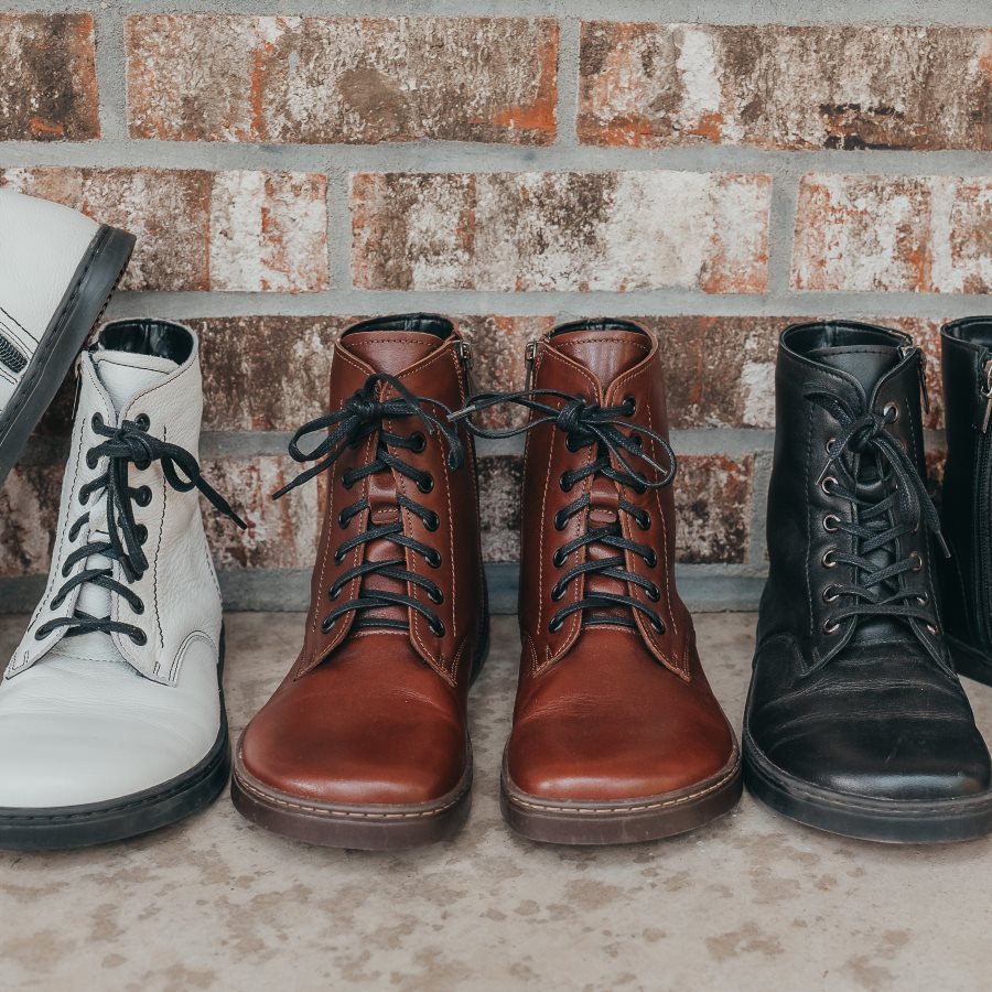 A photo of Peerko Go combat boots made with smooth leather, inside zippers, and rubber soles. Three pairs of boots are shown here in front of a brick wall. From right to left there are white boots with black soles/laces, cognac boots with black laces and brown soles, and all black boots. #color_cognac