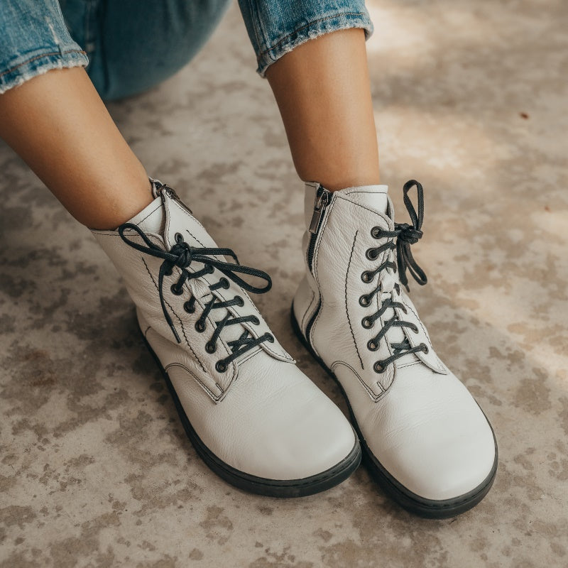 A photo of Peerko Go combat boots made with smooth leather and rubber soles. The boots are white in color, fleece lined, with a zipper at the side. Both boots are shown diagonally from the front right on a womans feet wearing loose blue jeans sitting on cement. #color_white