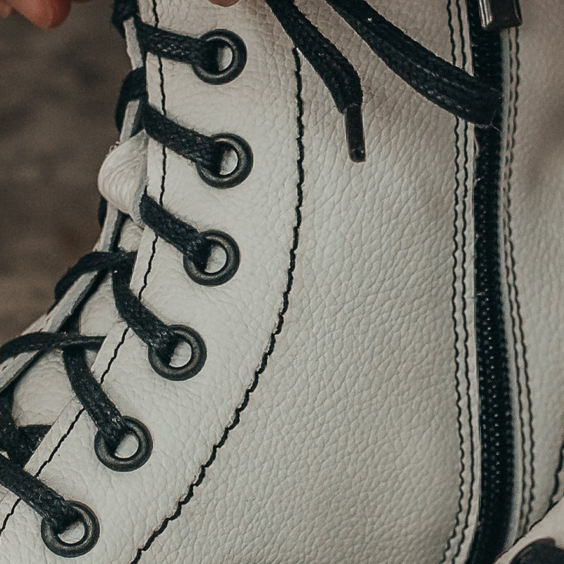 A photo of Peerko Go combat boots made with smooth leather and rubber soles. The boots are white in color, fleece lined, with a zipper at the side. This photo is a close up of the right boot's inside zipper and laces. Both of which are black. #color_white