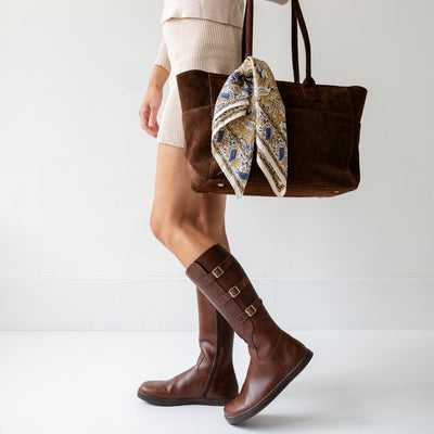 Side view of Peerko Joana brown knee-high boots worn with a cream outfit and brown suede tote bag. #color_brown