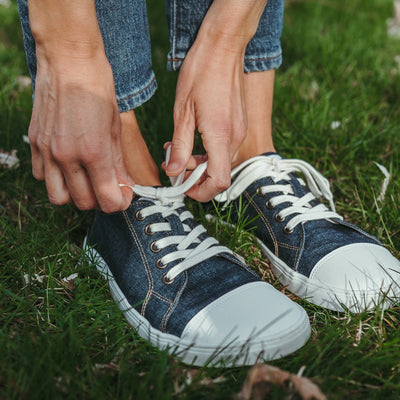 Dark Denim Peerko Origin sneakers with a white leather toe cap, soles, and laces. Shoes are shown on a woman wearing loose, cropped jeans standing in grass leaning over to tie the left lace. #color_denim