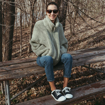 Dark Denim Peerko Origin sneakers with a white leather toe cap, soles, and laces. Shoes are shown facing diagonally right on a woman wearing loose, cropped jeans and a grey sherpa, zip-up sweater and sunglasses sitting on a weathered wooden picnic table. #color_denim