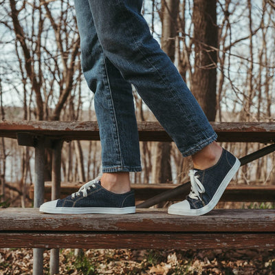 Dark Denim Peerko Origin sneakers with a white leather toe cap, soles, and laces. Shoes are shown facing left on a woman wearing loose, cropped jeans walking towards the left on a worn painted picnic bench. #color_denim