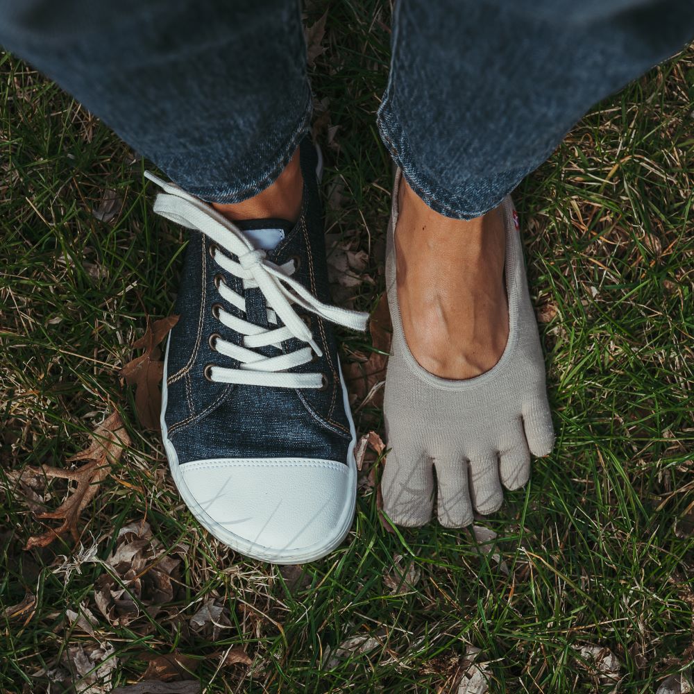 Dark Denim Peerko Origin sneakers with a white leather toe cap, soles, and laces. Right shoe is shown from above on a woman wearing loose, cropped jeans standing in grass. Left foot is wearing a no-show toe sock next to the right foot. #color_denim