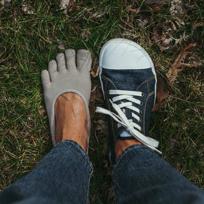 Dark Denim Peerko Origin sneakers with a white leather toe cap, soles, and laces. Right shoe is shown from above on a woman wearing loose, cropped jeans standing in grass. Left foot is wearing a no-show toe sock next to the right foot. #color_denim