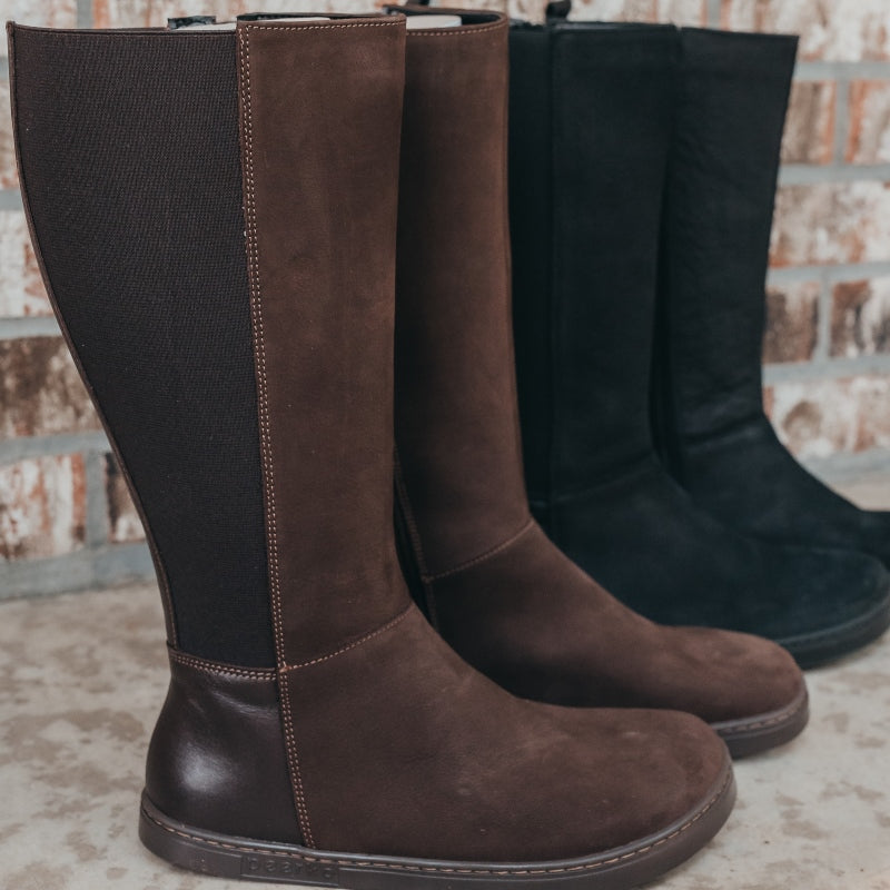 A photo of Peerko Regina riding boots made from smooth and suede leather and rubber soles. The boots are brown in color with a tall riding boot elastic paneled shaft, zippers, and lined with felt. Both boots are shown beside each other from the right against a brick background with the black boots beside them. #color_brown