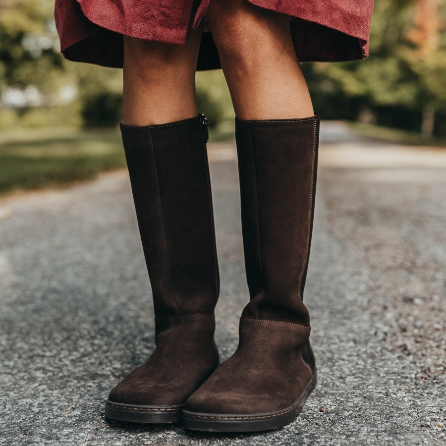 A photo of Peerko Regina riding boots made from smooth leather and rubber soles. The boots are brown in color with a tall riding boot elastic paneled shaft, zippers, and lined with felt. Both boots are shown on a woman's feet with a view of the knees down. The woman is wearing a red skirt and the boots and is standing on a paved road. #color_brown