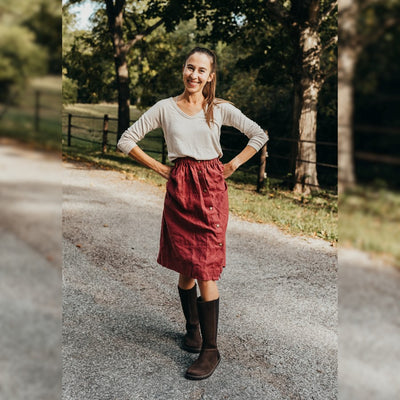 A photo of Peerko Regina riding boots made from smooth leather and rubber soles. The boots are brown in color with a tall riding boot elastic paneled shaft, zippers, and lined with felt. Both boots are shown on a woman's feet from the front. The woman is wearing a tan long sleeve shirt and a red skirt and is standing with her hands on her hips on a paved road. #color_brown