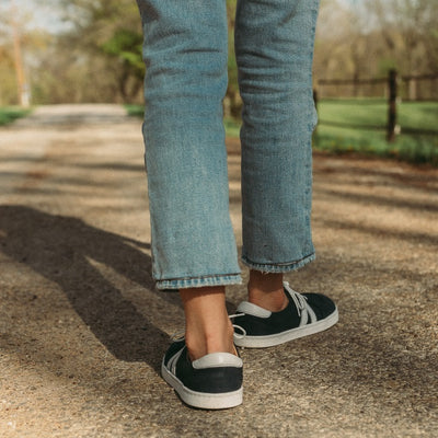 A photo of navy blue Peerko Street sneakers made with leather and recyclable soles. White pointed A-shaped detail present on both sides. Both shoes are shown from the back here on womens feet. Woman is standing on a gravel country road wearing lightwash ankle height jeans. #color_navy
