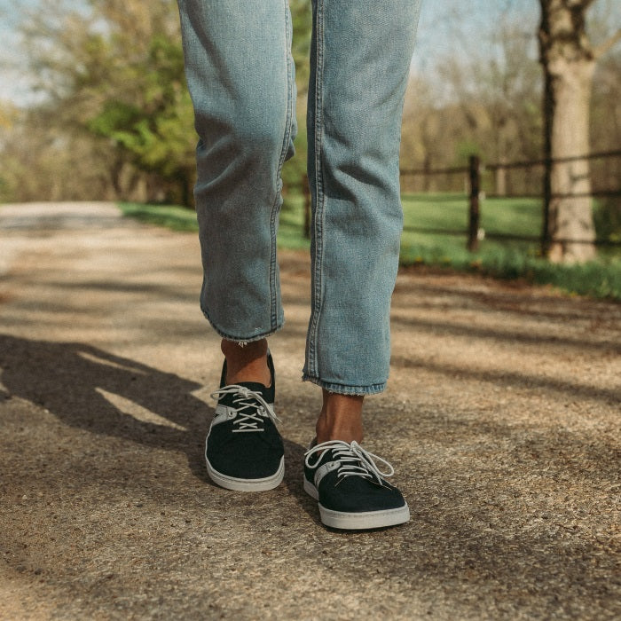 A photo of navy blue Peerko Street sneakers made with leather and recyclable soles. White pointed A-shaped detail present on both sides. Both shoes are shown from the front here on womens feet. Woman is standing on a gravel country road wearing lightwash ankle height jeans. . #color_navy
