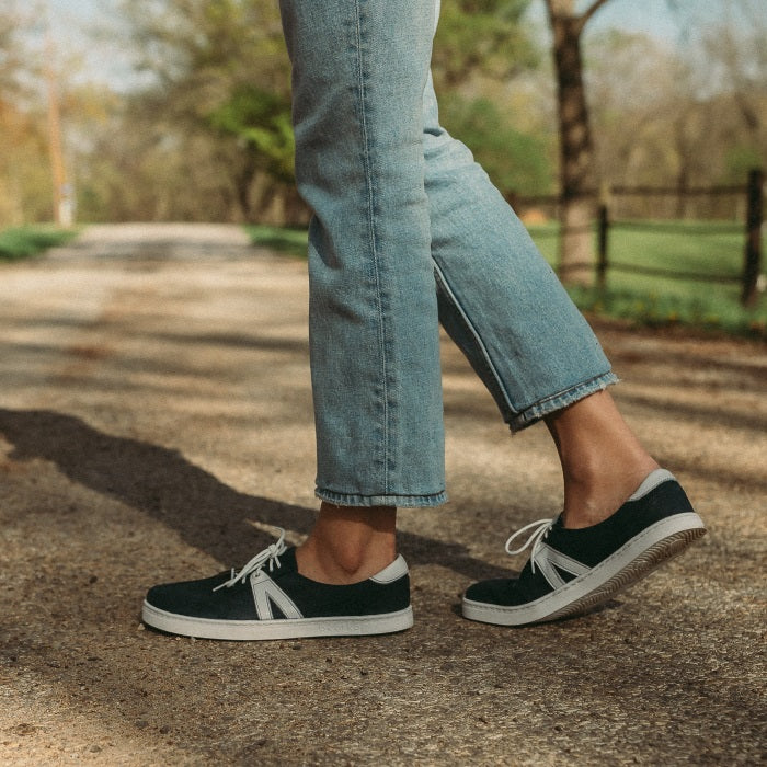 A photo of navy blue Peerko Street sneakers made with leather and recyclable soles. White pointed A-shaped detail present on both sides. Both shoes are shown from the left side here on womens feet with the right foot behind the left bent on the ground. Woman is standing on a gravel country road wearing lightwash ankle height jeans. . #color_navy