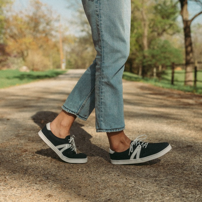 A photo of navy blue Peerko Street sneakers made with leather and recyclable soles. White pointed A-shaped detail present on both sides. Both shoes are shown from the right side here on womens feet with the left foot behind the right bent on the ground and the right foot resting on the heel. Woman is standing on a gravel country road wearing lightwash ankle height jeans. . #color_navy