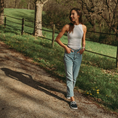 A photo of navy blue Peerko Street sneakers made with leather and recyclable soles. White pointed A-shaped detail present on both sides. Both shoes are shown from the front here on womens feet. Woman is standing on a gravel country road wearing lightwash ankle height jeans and  a white tank top looking to her right. #color_navy