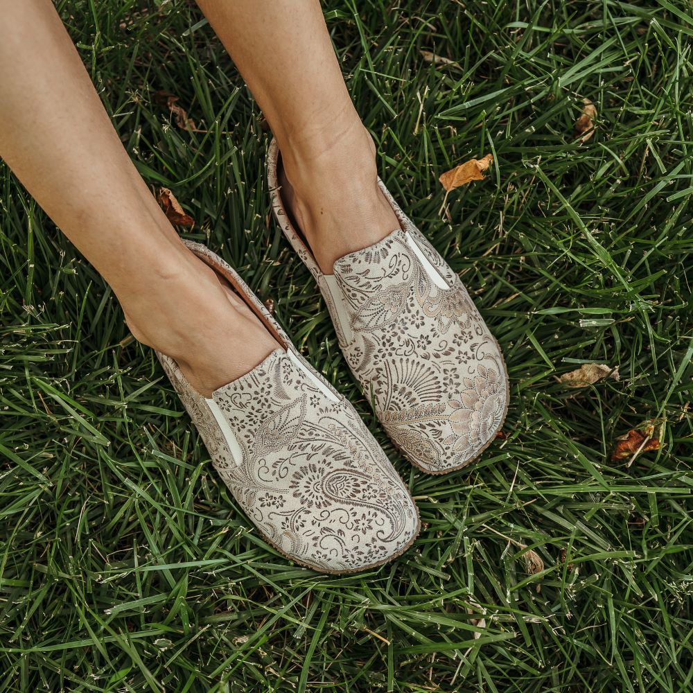 Light tan Peerko Trim leather loafer with a metallic floral pattern, tan rubber soles, and two small elastic pannels. Both shoes are shown from above on a tan woman sitting in grass facing diagonally right. #color_savana