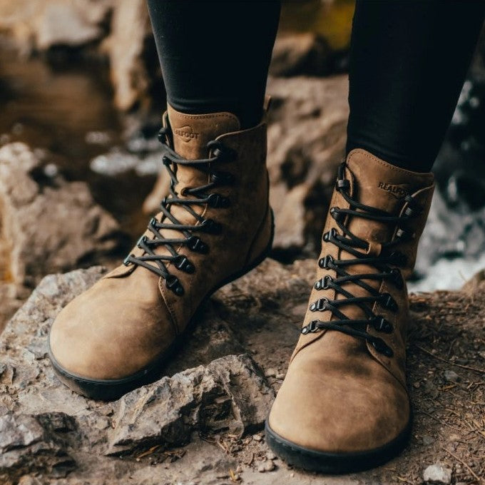 Light Brown shearling-lined leather Realfoot Farmer Winter boots with black laces, eyelets, and soles. Boots go over the ankle with padding and a pull tab in the back and a longer tongue. Two quick lace hooks are at the top of the laces and soles are stitched and glued. Both boots are shown from the front on a person wearing black leggings tucked into the boots standing on rocks. #color_light-brown