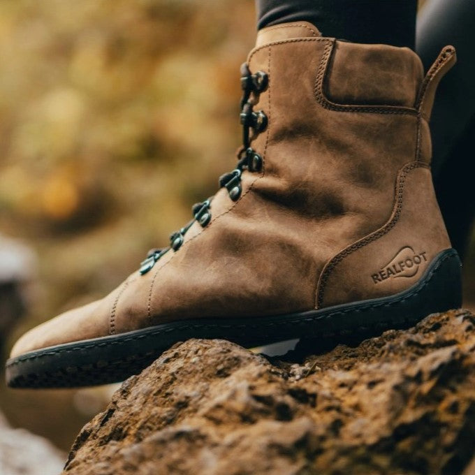 Light Brown shearling-lined leather Realfoot Farmer Winter boots with black laces, eyelets, and soles. Boots go over the ankle with padding and a pull tab in the back and a longer tongue. Two quick lace hooks are at the top of the laces and soles are stitched and glued. Left boot is shown facing left on a person wearing black leggings tucked into the boots standing on rocks. #color_light-brown