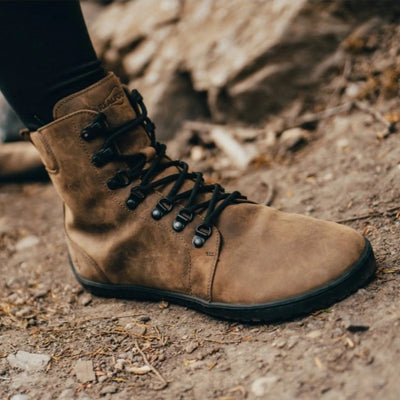 Light Brown shearling-lined leather Realfoot Farmer Winter boots with black laces, eyelets, and soles. Boots go over the ankle with padding and a pull tab in the back and a longer tongue. Two quick lace hooks are at the top of the laces and soles are stitched and glued. Right boot is shown facing diagonally right on a person wearing black leggings tucked into the boots standing on dirt. #color_light-brown