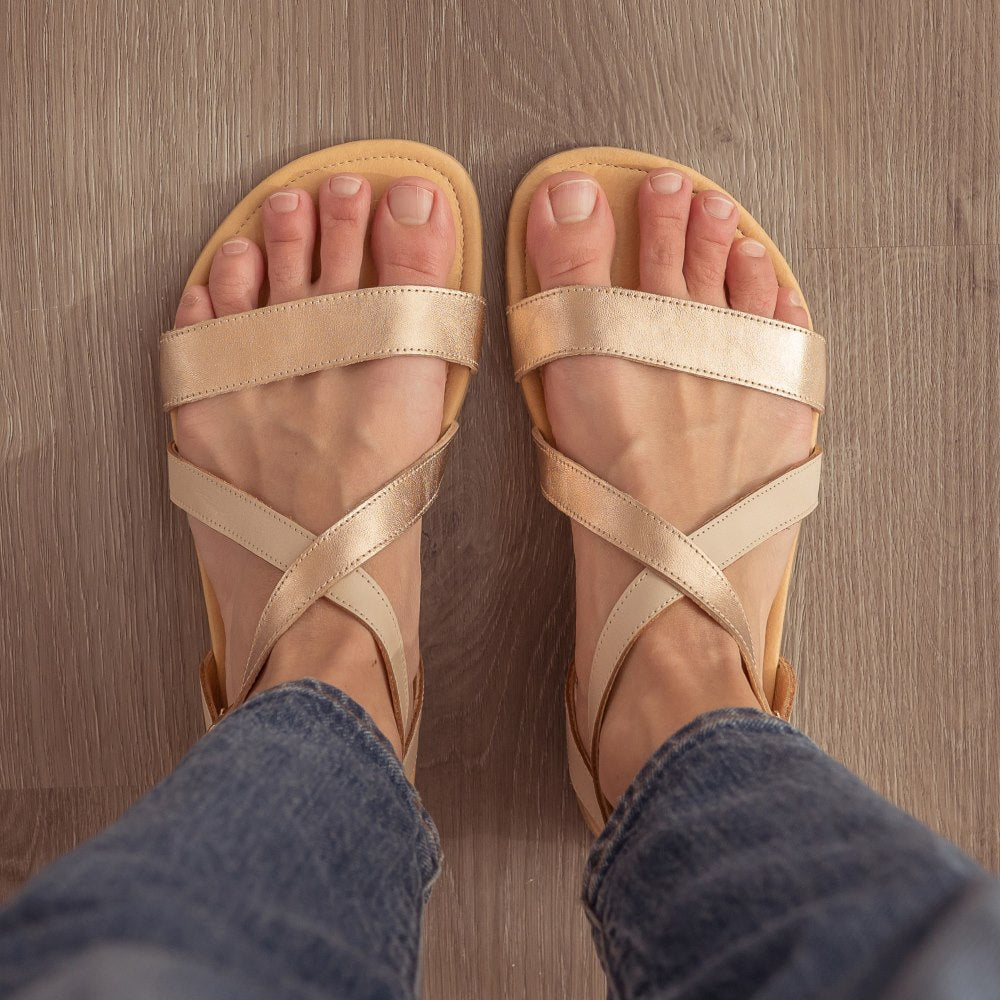 A photo of Rose Gold Shapen Calla Sandals made with leather and tan rubber soles. The sandals have a slight sparkle on the toe strap and one of the foot crossing straps. The other foot crossing strap is a creamy white. Both sandals are shown here from above here on a woman wearing medium-wash skinny jeans standing on a grey wooden floor. #color_rose-gold