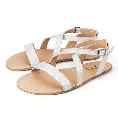 A photo of white Shapen Calla Sandals made with leather and tan rubber soles. The sandals have a slight sparkle on the toe strap and one of the foot crossing straps. The other foot crossing strap is a creamy white. Both sandals are shown from the left side beside each other against a white background in this photo. #color_white