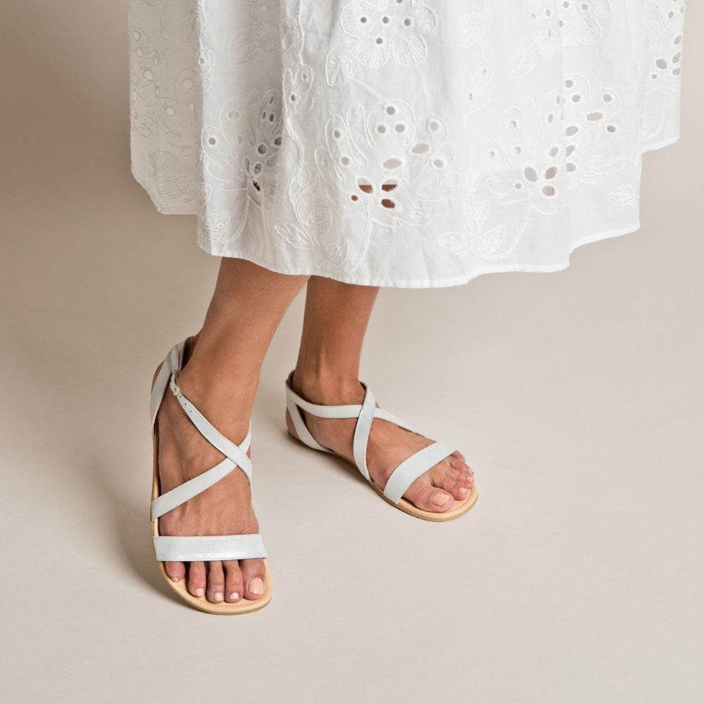 A photo of White Shapen Calla Sandals made with leather and tan rubber soles. The sandals have a slight sparkle. Left sandal is shown from the front right side against a cream background in this photo. Shown on a tan woman wearing a white lacey 3/4 length dress. #color_white