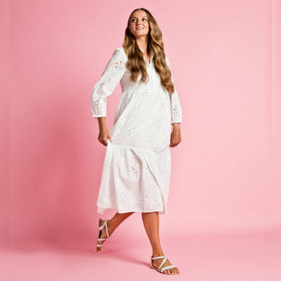 A photo of White Shapen Calla Sandals made with leather and tan rubber soles. The sandals have a slight sparkle. Left sandal is shown from the front right side against a pink background in this photo. Shown on a tan woman with long brown hair wearing a white lacey 3/4 length dress. #color_white