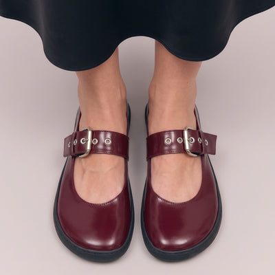 Front view of Shapen Coco bordeaux glossy Mary Jane shoes with wide silver buckle straps worn on a person’s feet against a soft neutral background. #color_bordeaux
