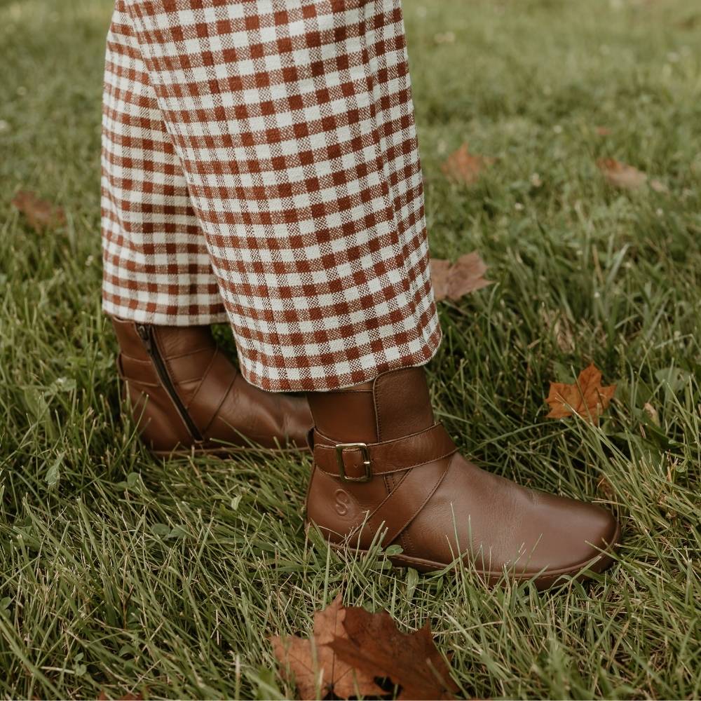 All brown leather Shapen Divine ankle boots with a pull tab, a wrap-style ankle strap, gold buckle, and a side zipper. Both boots are shown facing right on a woman wearing cropped/loose/white and brown gingham patterned pants standing in grass sprinkled with leaves. #color_brown