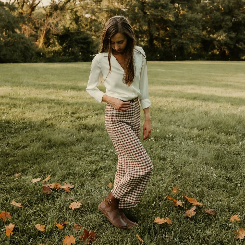 All brown leather Shapen Divine ankle boots with a pull tab, a wrap-style ankle strap, gold buckle, and a side zipper. Both boots are shown facing right on a light-skinned woman with long/straight/brown hair wearing a white collared blouse tucked into cropped/loose/white and brown gingham patterned pants standing in grass sprinkled with leaves. #color_brown