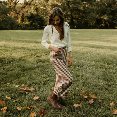 All brown leather Shapen Divine ankle boots with a pull tab, a wrap-style ankle strap, gold buckle, and a side zipper. Both boots are shown facing right on a light-skinned woman with long/straight/brown hair wearing a white collared blouse tucked into cropped/loose/white and brown gingham patterned pants standing in grass sprinkled with leaves. #color_brown